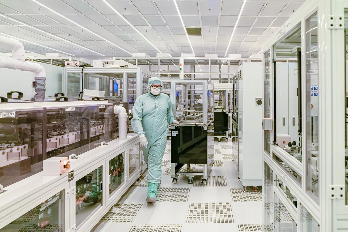 A technician in a cleanroom suit walking between semiconductor fabrication benches